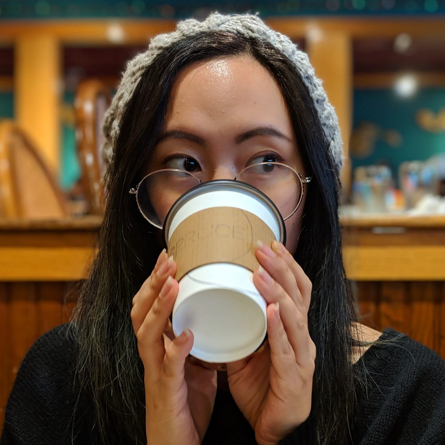 Portrait of Eileen drinking coffee. Girl with long black hair and glasses wearing a gray beanie, drinking from a paper coffee cup, with eyes looking to the right.