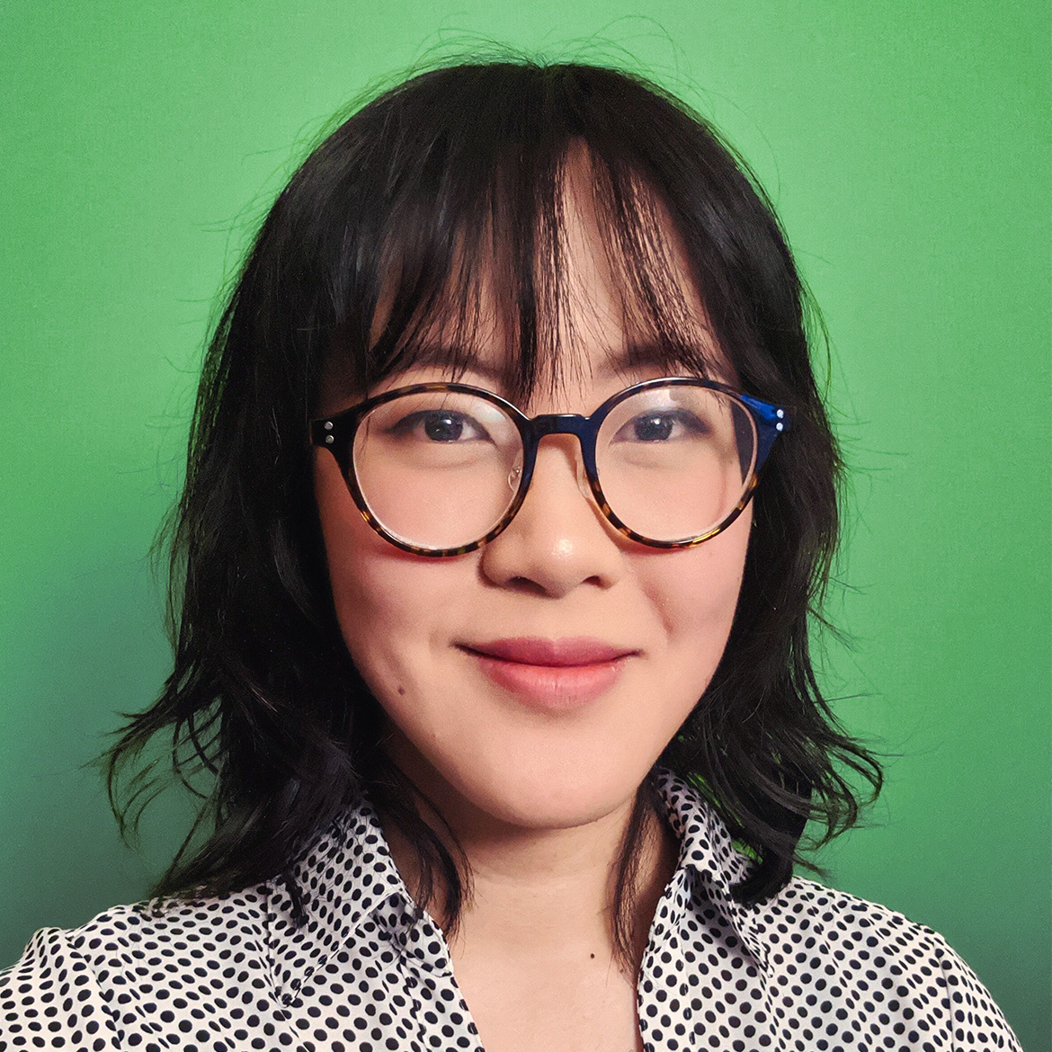 Headshot of Eileen smiling. Girl with black shoulder-length hair and bangs, round glasses, wearing a black and white polka dot shirt, in front of a green background.